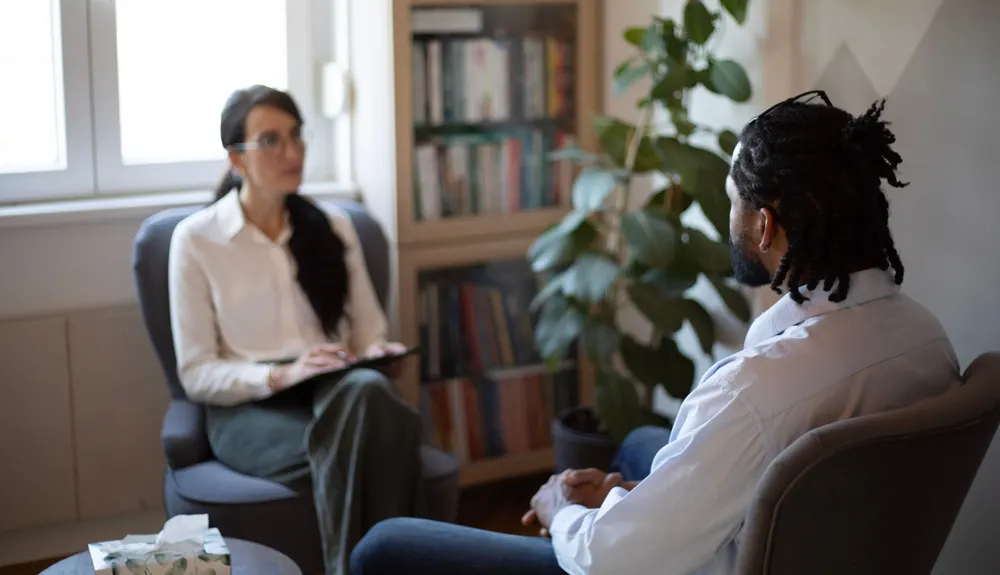 A counselor with a notepad sits in an armchair across from a bearded client in a book-lined office