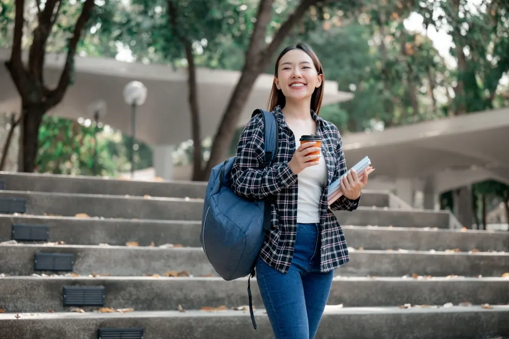 A smiling student carrying a backpack, notebooks and a coffee cup walks down wide campus steps