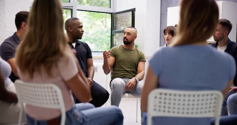 Group members seated in a circle listen as a man in an olive t-shirt speaks during a support group meeting