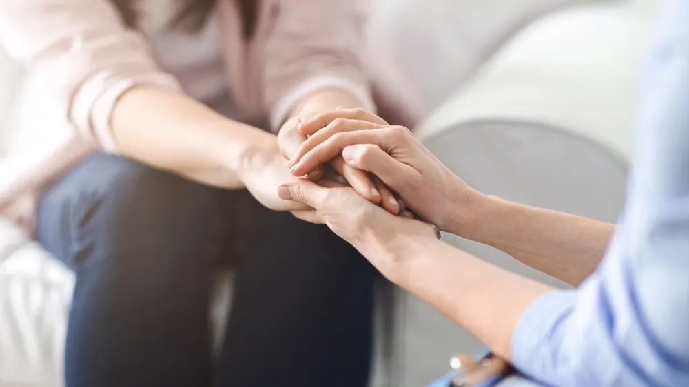 A close-up of a therapist in a light blue shirt holding a seated client's clasped hands during a session