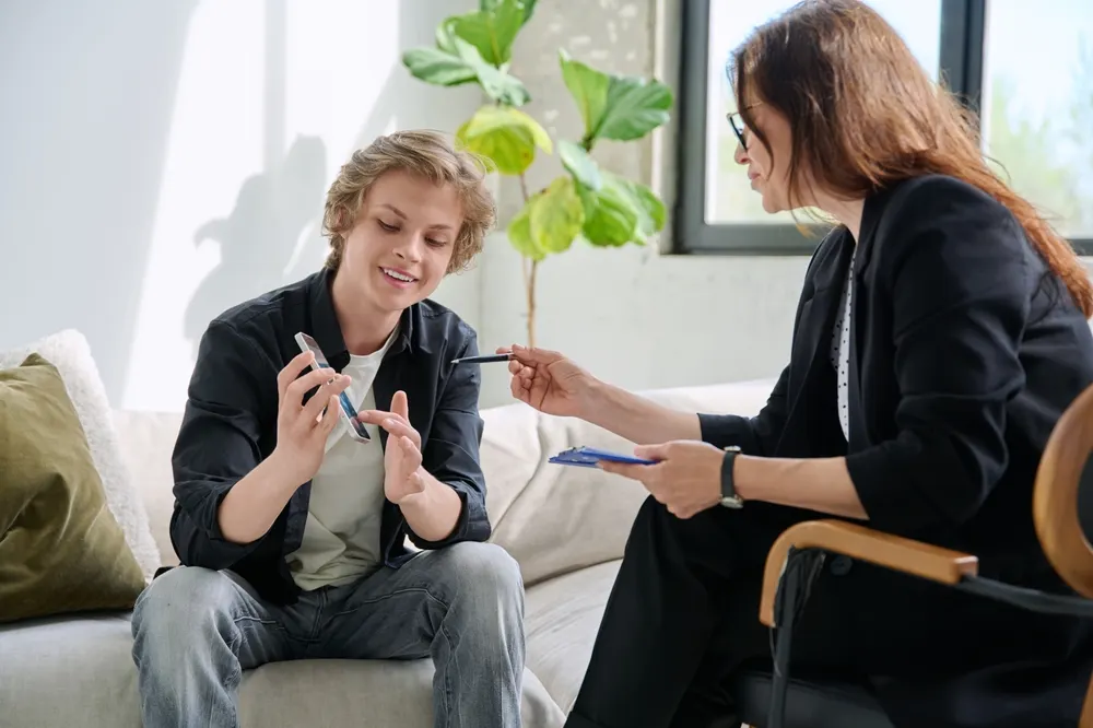A teenage boy gestures while talking on a sofa as a counselor in a black blazer holding a blue clipboard listens