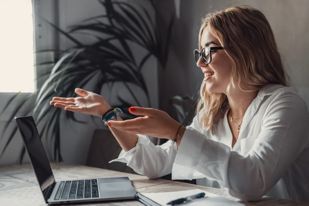 A woman in glasses and a white shirt gestures with both hands while speaking during a video call on her laptop