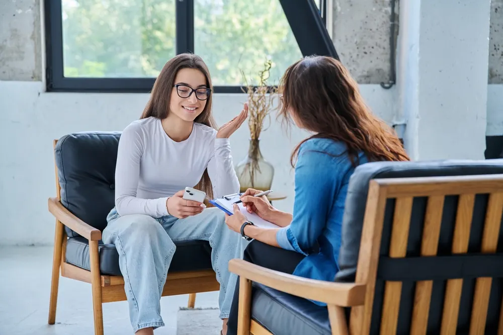 A counselor writing on a clipboard listens to a smiling young client during a counseling session