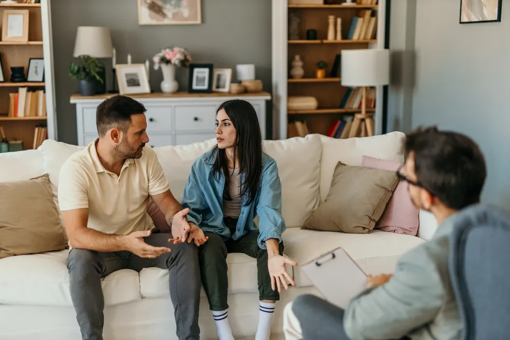 A couple sitting on a cream sofa gesture mid-conversation while a therapist with a clipboard listens in the foreground